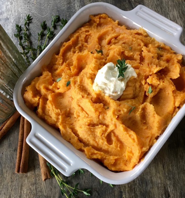 A photo of creamy mashed sweet potatoes in a casserole dish with butter and herbs on top.