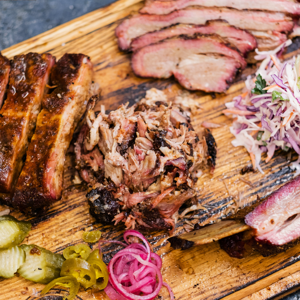 Wooden cutting board with 3 types of BBQ sliced meats as well as slaw, pickles, jalepeno peppers and red onion resting on it.