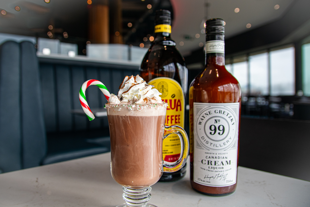 Photo of Hot Chocolate with a candy cane in the cup with 2 liquor bottles behind the cup.