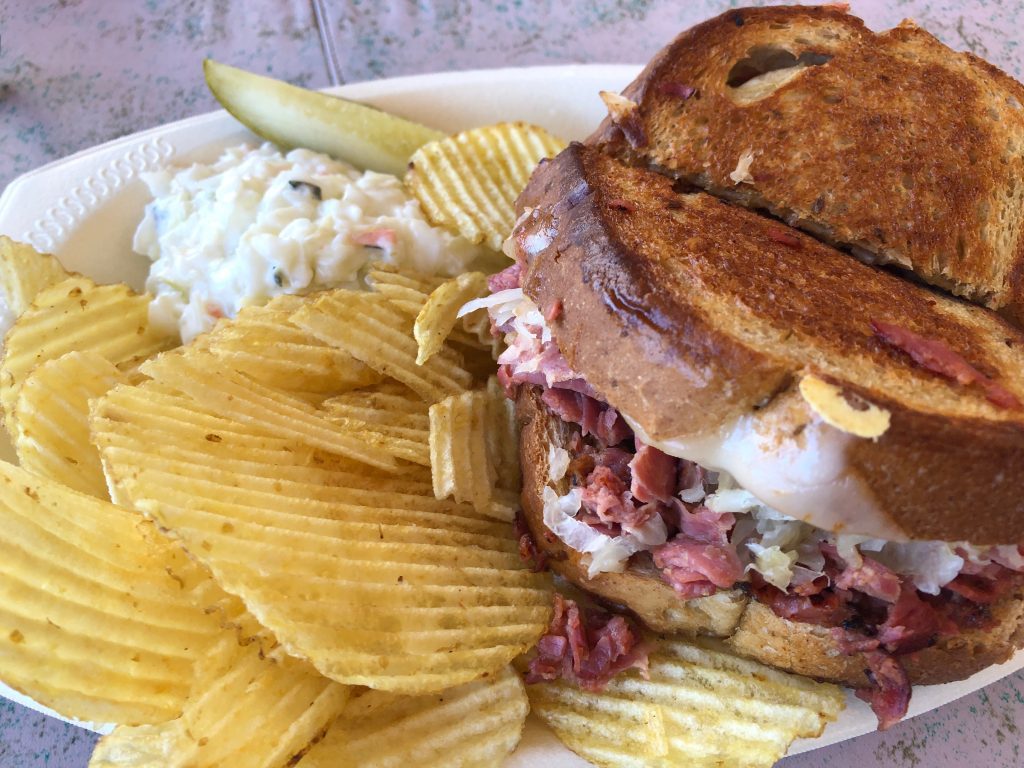 Photo of a sandwich wit a side of chips, pickle and slaw.