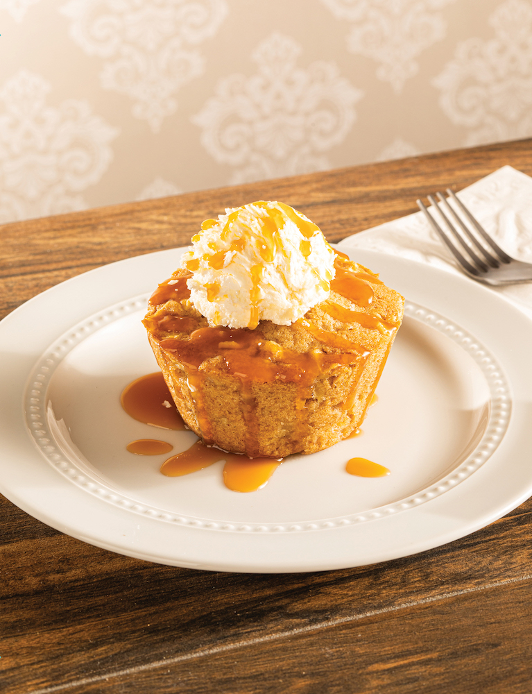 Johnny Appleseed's warm apple cake topped with vanilla ice cream and carmel drizzle on a whitle plate sitting on a wooden table.