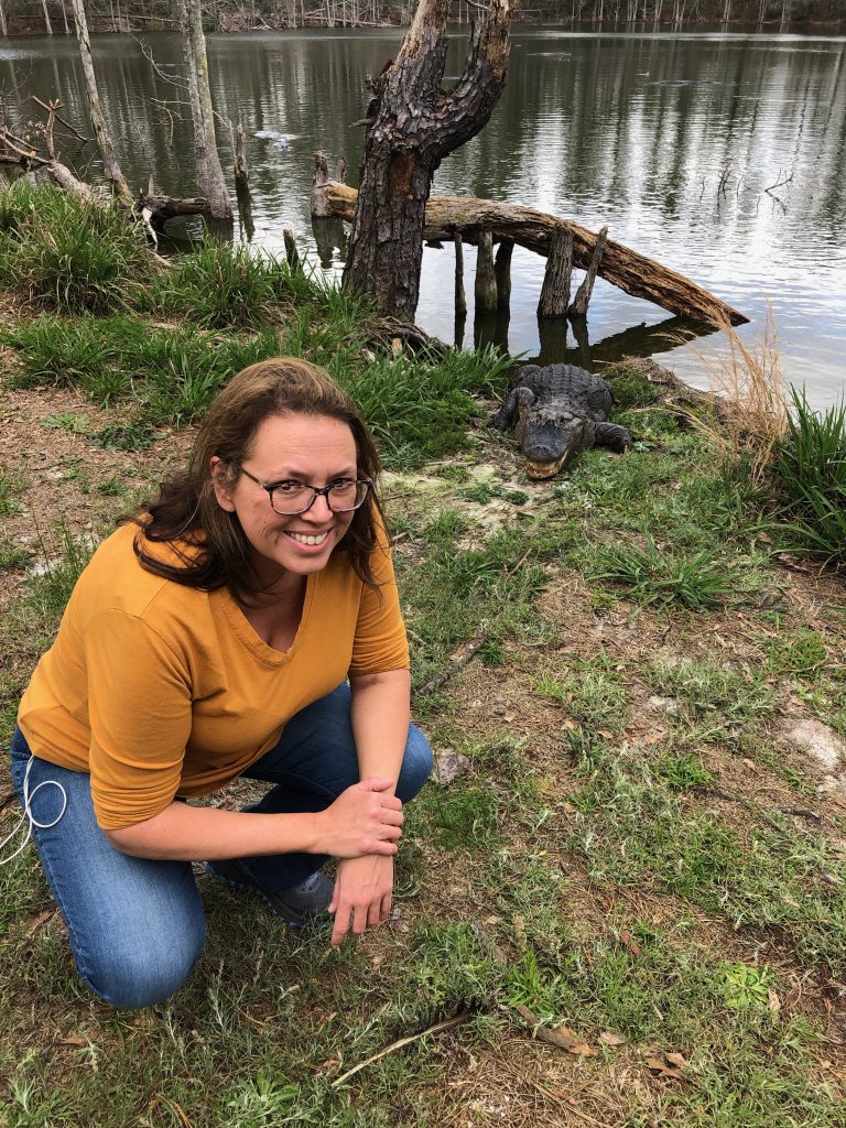 Photo of a women with an alligator behind her near the river.