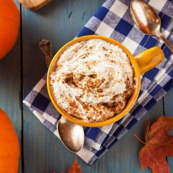 cup of pumpkin spice latte with whipped topping sprinkled with cinnamon set on a blue and white checkered napkin with a silver spoon resting next to it.