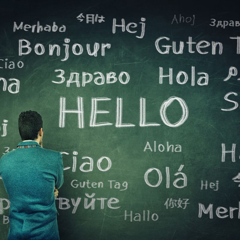 Green board with the word hello written in different languages with a person standing in front of it.